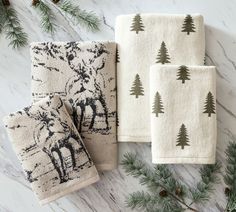 three hand towels with moose and pine trees printed on them next to evergreen branches, fir cones and pine cones