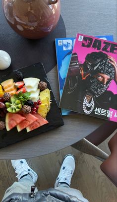 there is a plate of fruit and magazines on the table with someone's feet