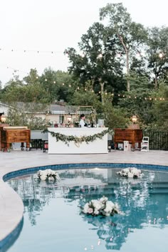 an outdoor wedding ceremony with flowers on the edge of the pool and lights hanging from the trees