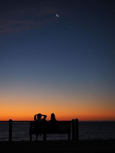 two people sitting on a bench watching the sun go down over the ocean at dusk