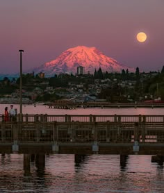 the full moon is setting over a mountain range in the distance as people stand on a pier