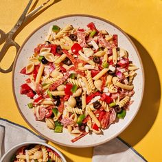 a bowl of pasta salad with olives, tomatoes, and other toppings next to a pair of scissors