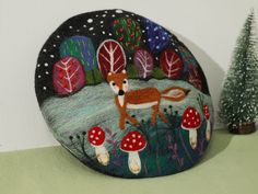 a painted rock with a deer and mushrooms in the forest next to a small christmas tree