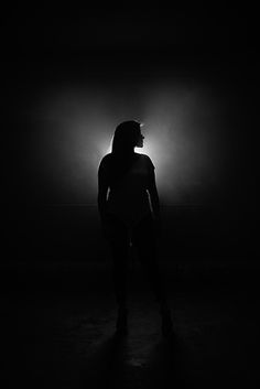 a woman standing in the dark with her back turned to the camera and light behind her