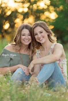 two women are sitting in the grass and posing for a photo with their arms around each other