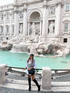 a woman standing in front of a fountain with statues on it's sides and wearing knee high boots