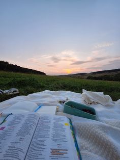 an open book sitting on top of a blanket in front of a field at sunset