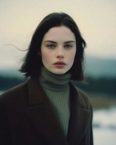 a woman with dark hair wearing a green turtle neck sweater and black coat looking at the camera