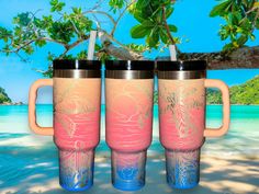 three travel mugs sitting next to each other in front of a tree on the beach