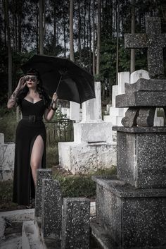 a woman in a black dress holding an umbrella over her head while standing next to some headstones