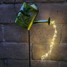 a green watering can with fairy lights hanging from it's side next to a brick wall