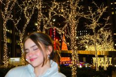 a woman standing in front of trees covered with christmas lights and smiling at the camera