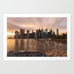 a city skyline is shown with water and rocks in the foreground, as the sun sets