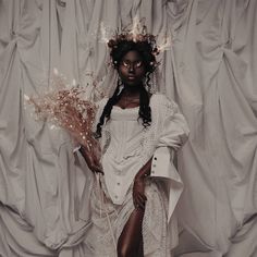 a woman in white dress standing next to a curtain with flowers on her head and legs