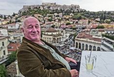 a man sitting at a table with a drink in front of him and a view of the city behind him