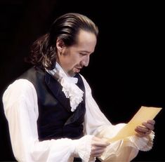 a man with long hair wearing a vest and holding a piece of paper in his hands