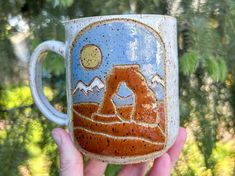 a hand holding up a ceramic mug with mountains and trees in the background