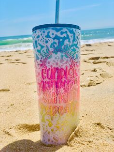 a colorful cup sitting on top of a sandy beach next to the ocean with words painted on it