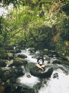 a man sitting on top of a rock in the middle of a river surrounded by trees