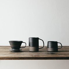 three black coffee cups sitting on top of a wooden table next to a white wall