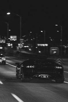 black and white photograph of cars driving down the road at night time with city lights in background