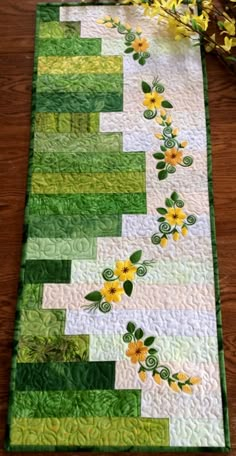 a quilted table runner with yellow flowers and green leaves on it, sitting on a wooden surface