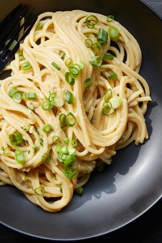 a black plate with noodles and green onions on it next to a knife and fork