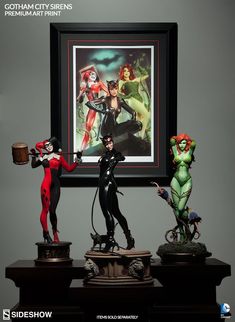 three statues are on display in front of a framed painting with the image of harley and catwoman