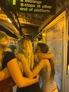 three girls hugging each other on a train platform at night time, with the sign in the background