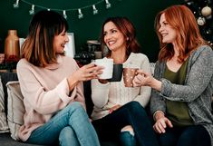 three women sitting on a couch holding coffee cups in their hands and smiling at each other