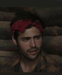 a man with a red bandana on his head looking at the camera while standing in front of logs