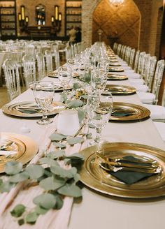 the table is set with gold plates and place settings, along with greenery on each plate