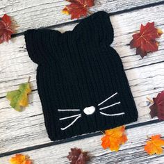 a black knitted cat hat with white eyes and ears on top of autumn leaves