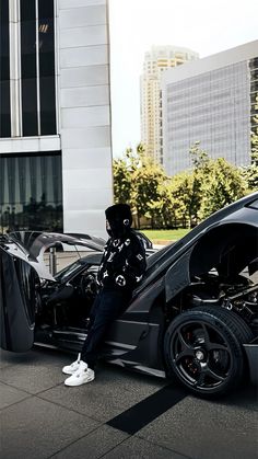 a man sitting on top of a black car next to a tall building in the city