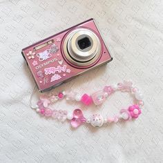 a pink camera sitting on top of a white bed next to a pink beaded bracelet