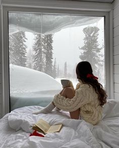 a woman sitting on top of a bed in front of a window with snow covered trees