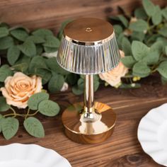 a gold table lamp sitting on top of a wooden table next to plates and flowers