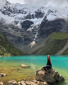 a woman sitting on top of a large rock next to a lake with snow covered mountains in the background