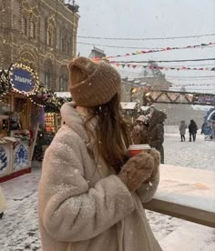 a woman standing in the snow holding a cup and looking at buildings with christmas lights on them