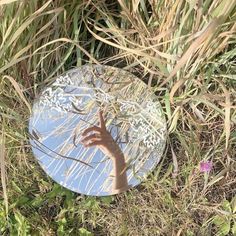 a round mirror sitting in the grass with a hand reaching up to it's reflection