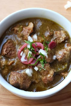 a white bowl filled with meat and vegetables on top of a wooden table next to a spoon