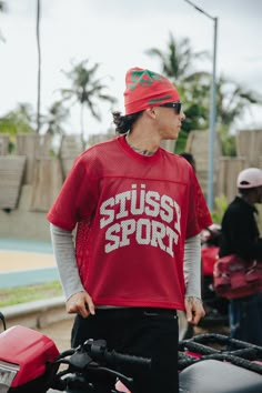 a man wearing a red shirt and hat standing next to a motorcycle with other people in the background