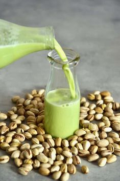 a green liquid being poured into a glass bottle surrounded by nuts and pistachio