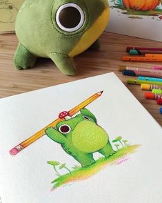 a green frog with a pencil in its mouth sitting next to some colored crayons