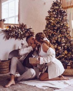 a man and woman sitting on the floor kissing in front of a decorated christmas tree