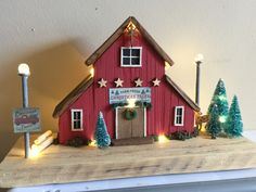 a small red barn with christmas lights on the roof and trees around it in front of a white wall