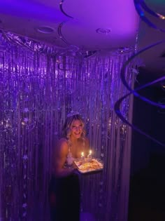 a woman holding a cake with lit candles in her hand while standing next to a purple curtain