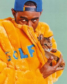 a man holding a cat in his right hand while wearing a blue hat and yellow jacket