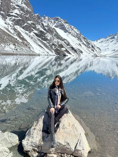 a woman sitting on top of a rock next to a body of water with snow covered mountains in the background