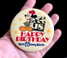 a hand holding a mickey mouse button with the words happy birthday written in red on it